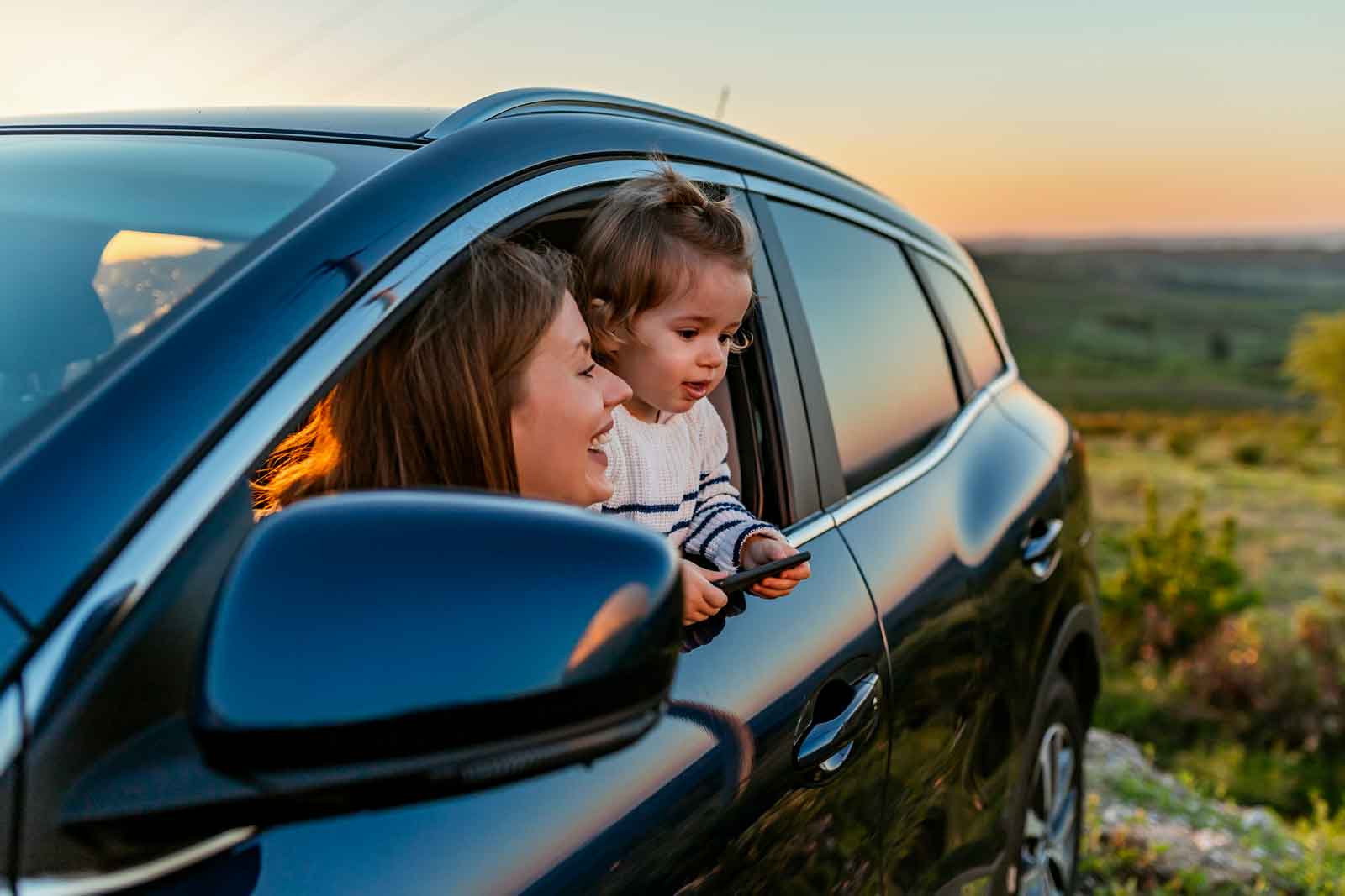 Family vacation theme. Young woman with kid looking out from car window during a road trip in summer. Photo of happy Caucasian mother and son sticking heads out of the window of a car looking at the scenery.