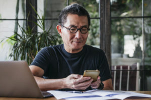 Asian man in his 50's working remotely, using a mobile phone to process technology transactions