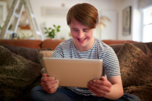 Young Downs Syndrome Man Sitting On Sofa Using Digital Tablet At Home