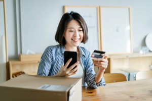 Cheerful young woman shopping online with smartphone and making mobile payment with credit card on hand, receiving a parcel by home delivery service. Technology makes life so much easier