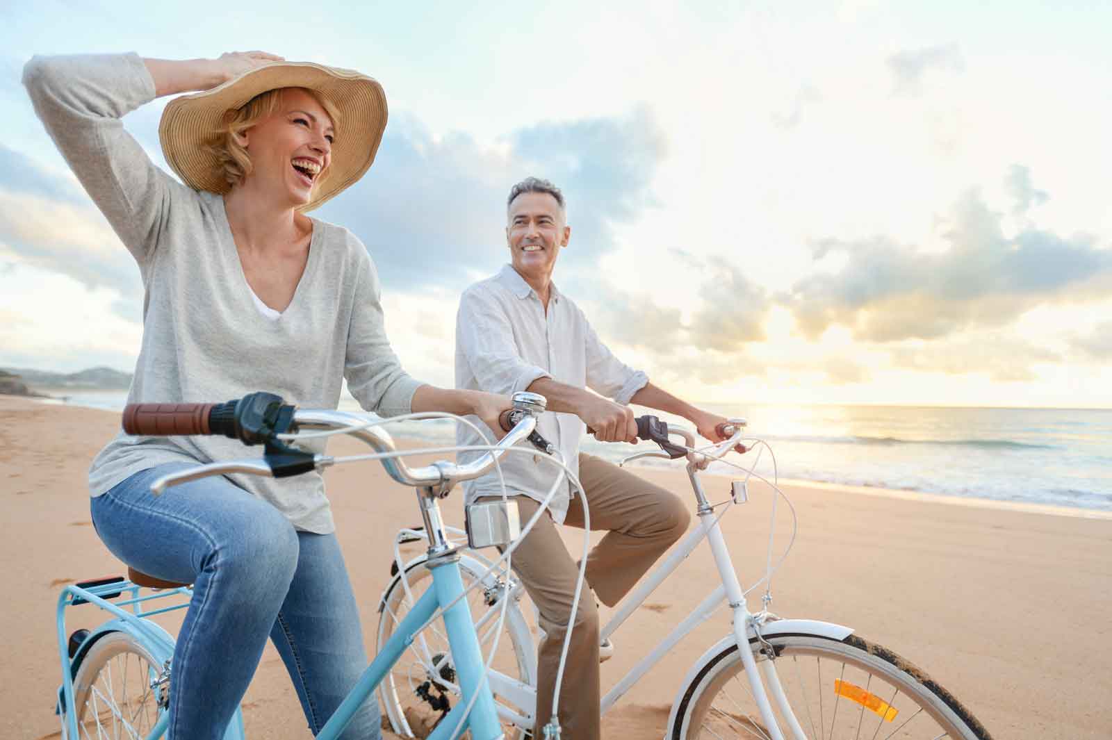 Mature couple cycling on the beach at sunset or sunrise. They are laughing and having fun. They are casually dressed. Could be a retirement vacation.