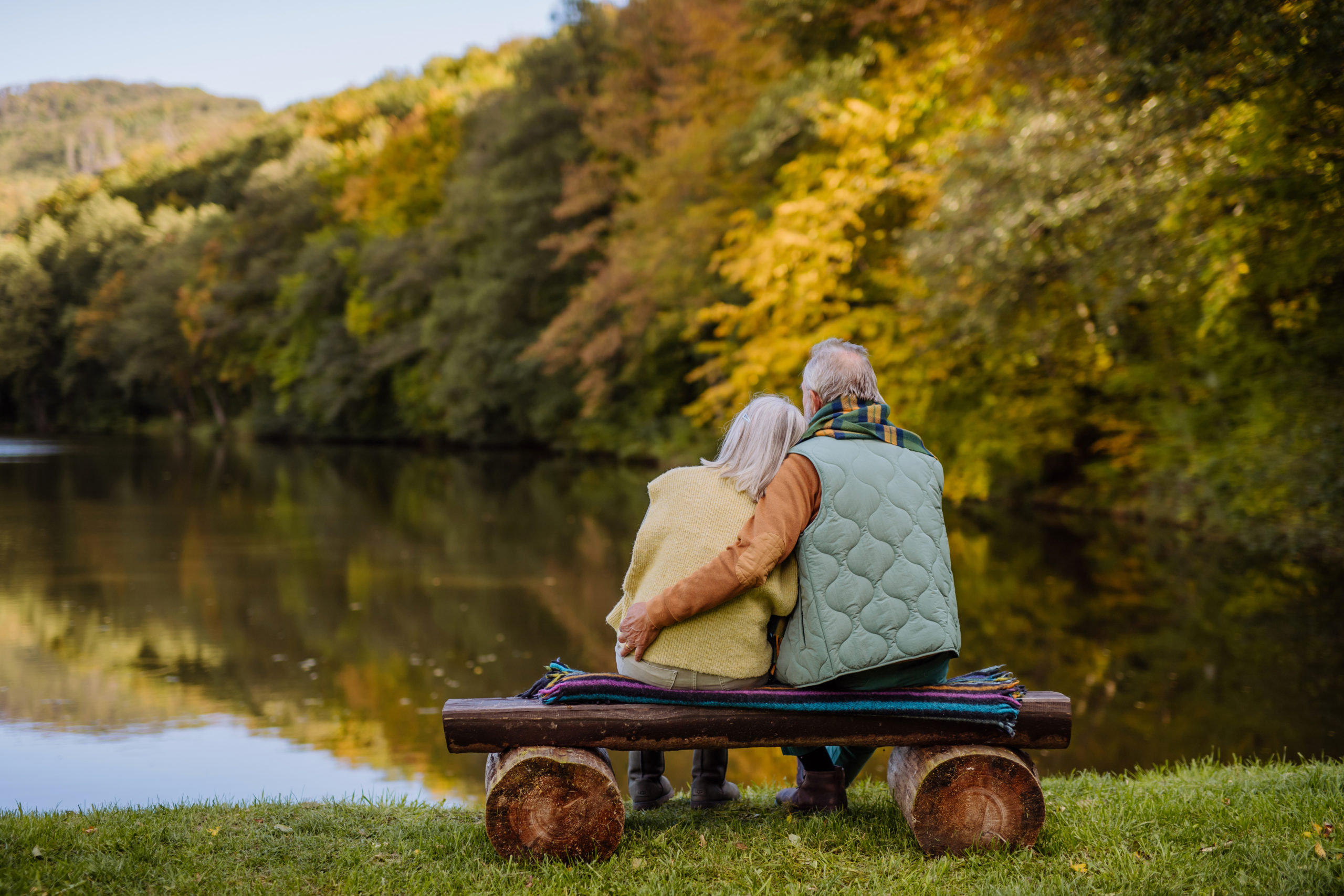 Senior couple in love sitting together on a bench looking at lake, during autumn day. Rear view.