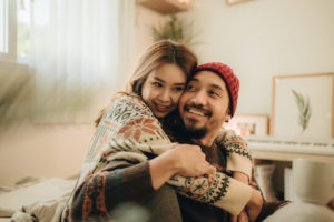 Happy young family couple relaxing talking laughing holding cups drinking coffee tea sitting on bed together in bedroom, loving husband and wife bonding enjoying pleasant conversation at home