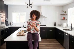 Woman At Home Sitting On Kitchen Island Whilst Using Mobile Phone