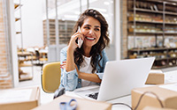 Happy young businesswoman speaking on the phone while working in a warehouse. Online store owner making plans for product shipping. Creative female entrepreneur running an e-commerce small business.