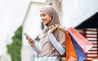 New mobile app for shopaholics. Happy arab woman in headscarf with shopping bags using smartphone, standing on street, side view, free space
