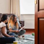 Two women going through personal finances at home sitting on the floor in the lounge looking down at a receipt. They are smiling at each other positively. Shot through a doorway