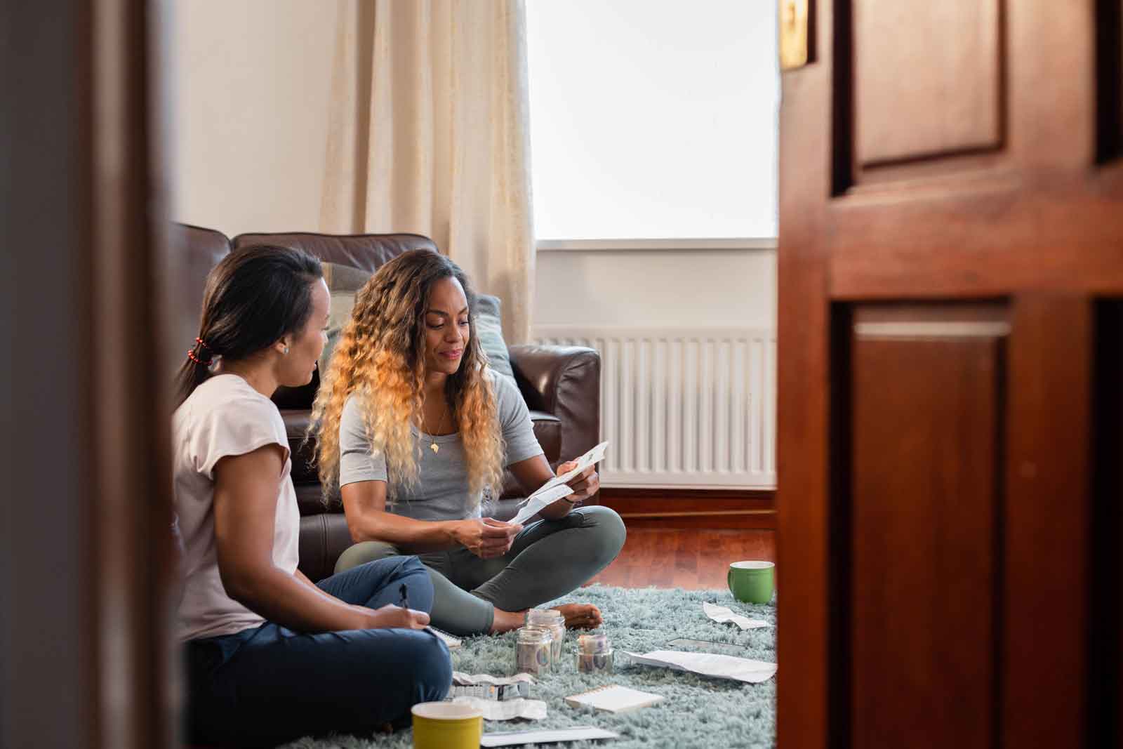 Two women going through personal finances at home sitting on the floor in the lounge looking down at a receipt. They are smiling at each other positively. Shot through a doorway