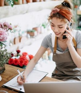 Young Asian Female Florist, Owner Of Small Business Flower Shop, Talking On Smartphone While Working On Digital Tablet In Workplace. Checking Stocks, Taking Customer Orders, Selling Products Online. Daily Routine Of Running Small Business With Technology