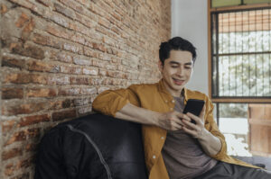 A young smiling Asian man chatting on his smartphone and sitting in the living room.