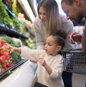 A pre-school age girl helps her parents pick out veggies in the produce section at the grocery store. She is reaching for a red bell pepper. Her mother is Caucasian and her father is African-American.