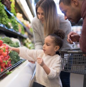 A pre-school age girl helps her parents pick out veggies in the produce section at the grocery store. She is reaching for a red bell pepper. Her mother is Caucasian and her father is African-American.
