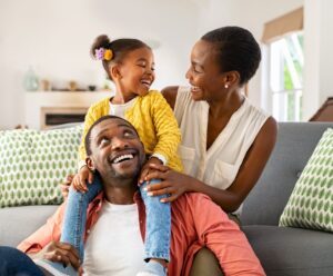 Cheerful little girl sitting on father shoulder while playing with mother at home. Happy black family enjoying weekend at home. Cute little daughter sitting on fathers shoulder and play with her mom.