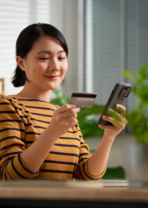 Young woman wearing striped shirt holding a card and a phone in her home office