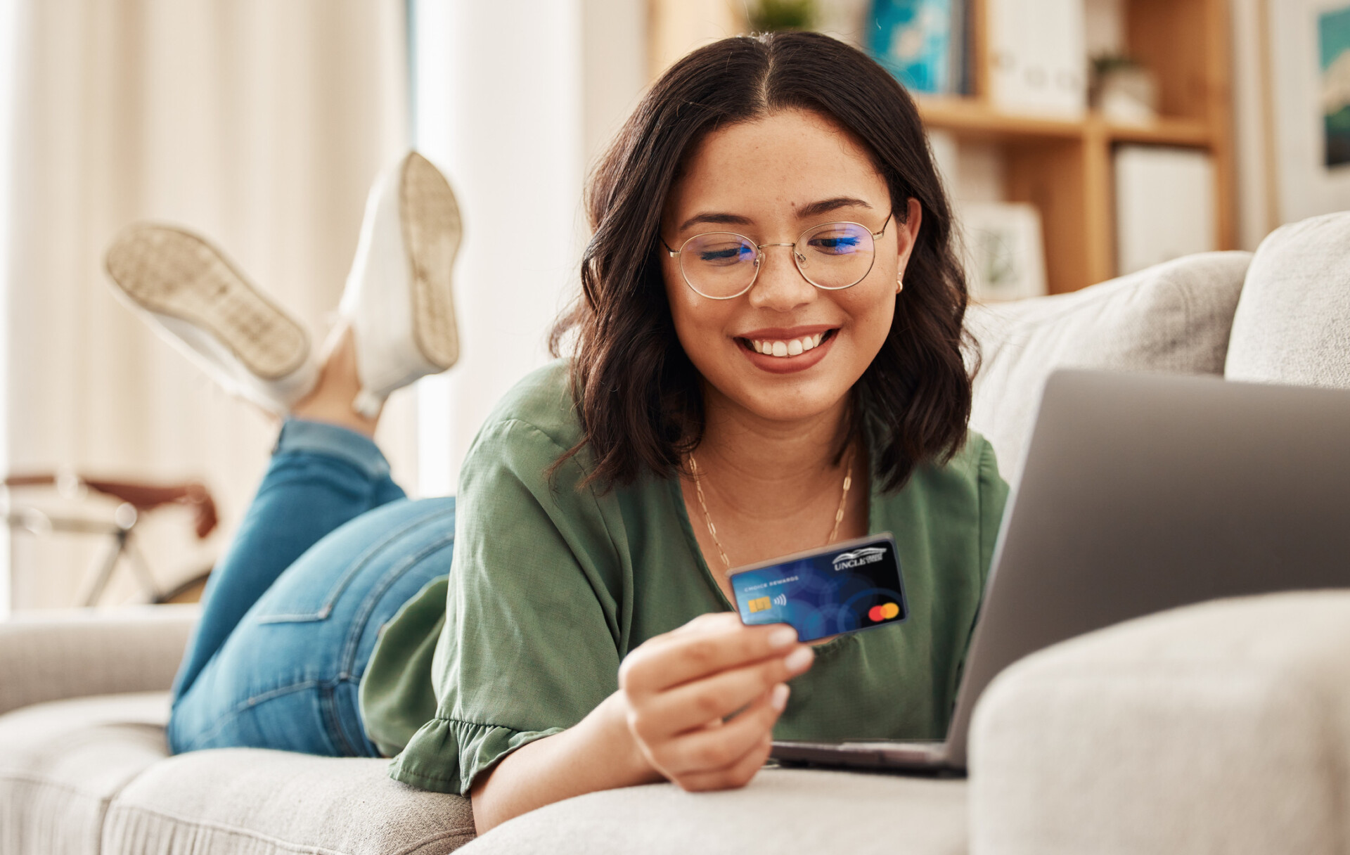 Woman lying on couch with laptop while happily looking at credit card.