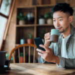 Young Asian Man Managing Online Banking With Mobile App On Smartphone, Taking Care Of His Money And Finances While Relaxing At Home. Banking With Technology