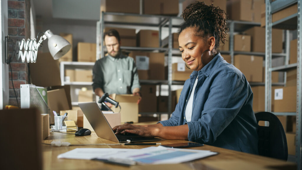 Diverse Male And Female Warehouse Inventory Managers Talking, Using Laptop Computer And Checking Retail Stock. Rows Of Shelves Full Of Cardboard Box Packages In The Background.