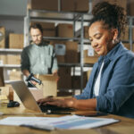 Diverse Male And Female Warehouse Inventory Managers Talking, Using Laptop Computer And Checking Retail Stock. Rows Of Shelves Full Of Cardboard Box Packages In The Background.