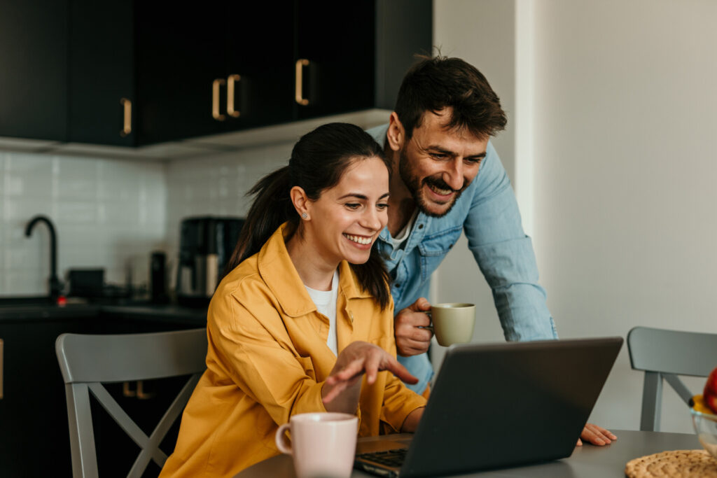 Happy Young Couple Husband And Wife Using A Laptop Computer Looking At A Screen Paying Bills Online, Planning Budget Discussing Finances Sit At The Home Table
