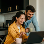 Happy Young Couple Husband And Wife Using A Laptop Computer Looking At A Screen Paying Bills Online, Planning Budget Discussing Finances Sit At The Home Table