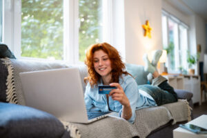 Young redhead Caucasian woman using credit card for online shopping, while relaxing on the sofa