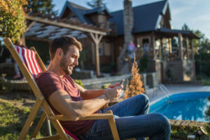 Happy Man Using Smart Phone In Deck Chair At The Back Yard.