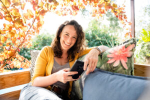 Portrait of a laughing woman with a smart phone relaxing outside on her patio sofa.