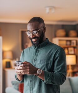 Happy man standing in his living room, smiling while using phone.