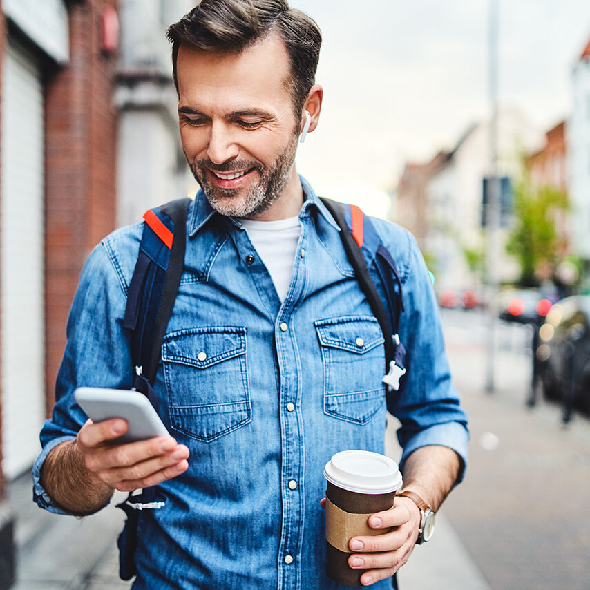 Man using phone with wireless headphones walking in the city and drinking coffee